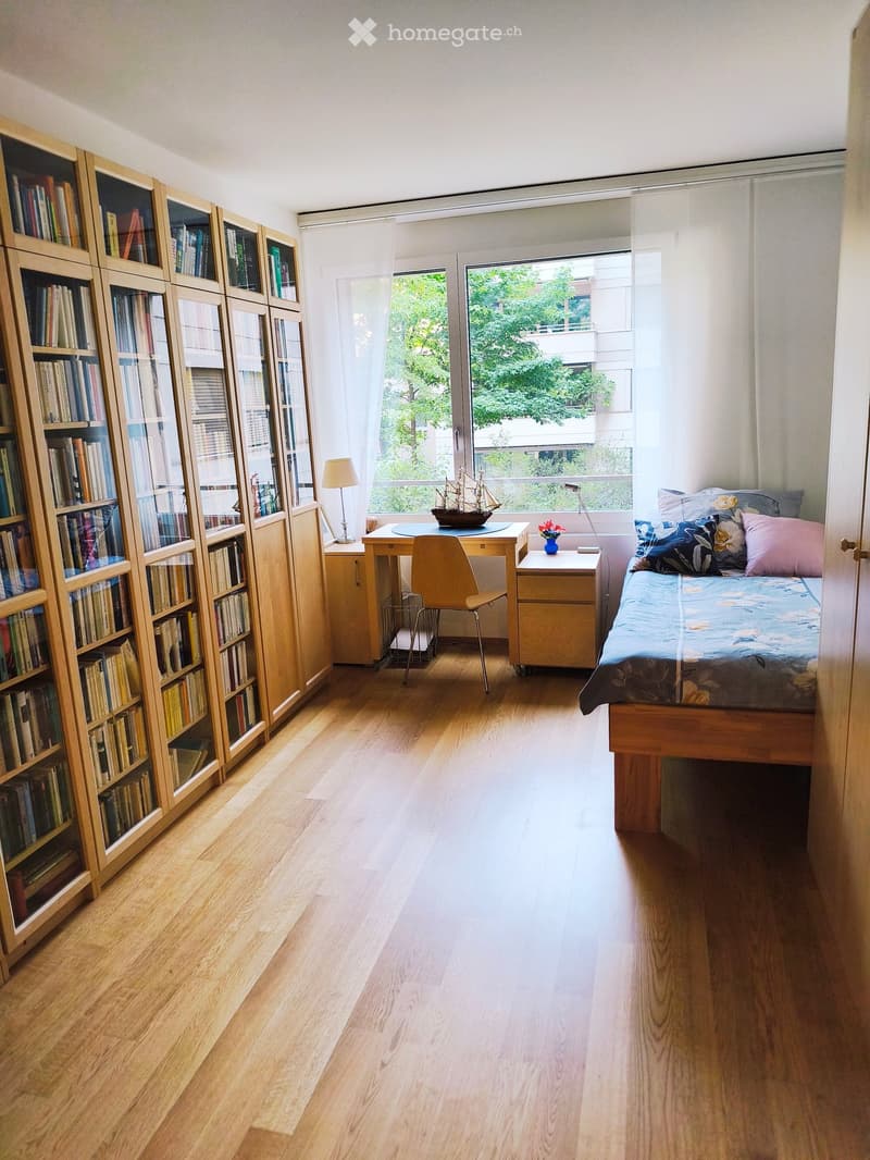 Bright, minimalist bedroom with wooden flooring, large window, bed with colorful bedding, bookshelf filled with books, desk with chair, and small nightstand with lamp and vase.