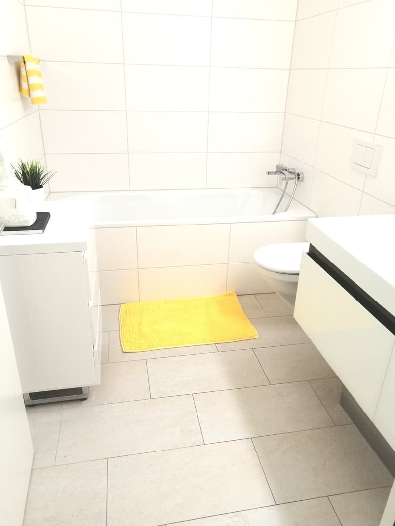 Bathroom with white tile walls, bathtub, toilet, white counters, yellow rug, cabinet with a small potted plant