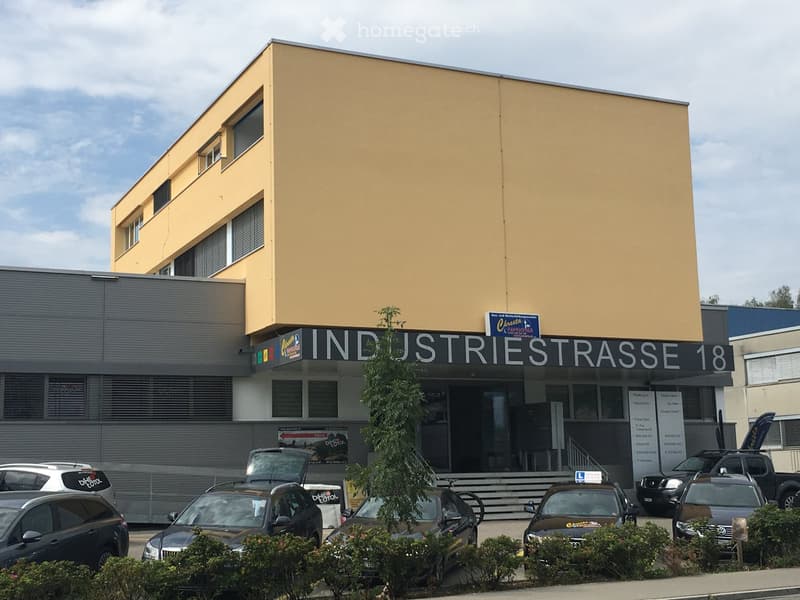 Two-story building with a yellow facade and gray lower section, labeled 'INDUSTRIESTRASSE 18', with several parked cars in front and a small tree near the entrance.