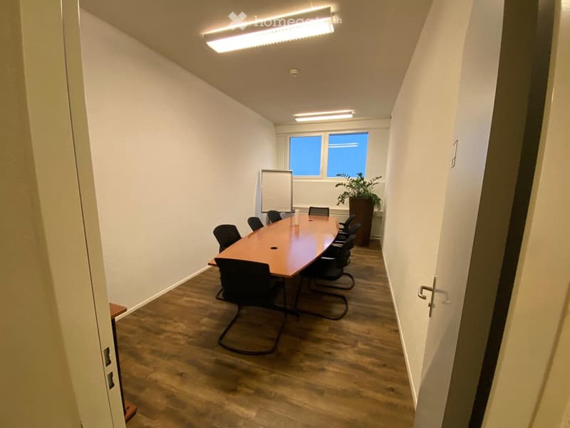 A conference room with a long wooden table, black chairs, a whiteboard, and a potted plant near the window.