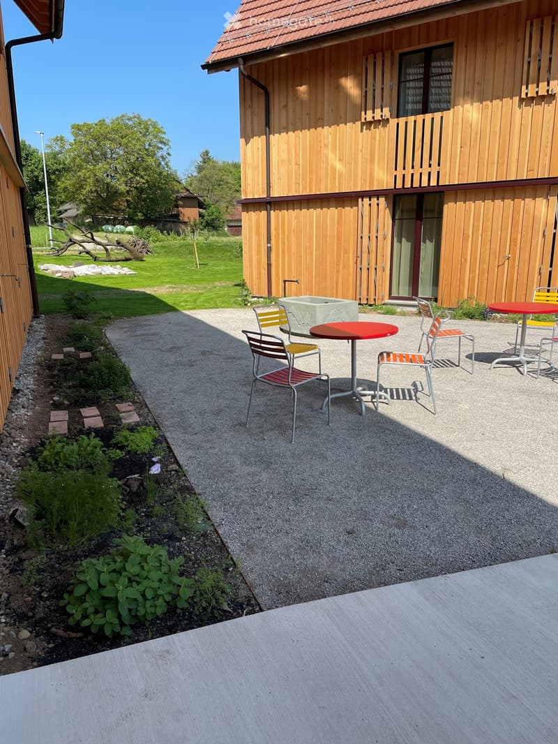 Outdoor patio area with a red table and yellow chairs, surrounded by a wooden house and garden.