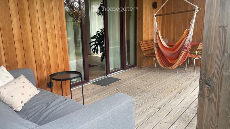 Wooden deck with a grey sofa, small round table, wooden chair, and hammock. Large glass doors lead to the interior, with indoor plants visible through the glass.