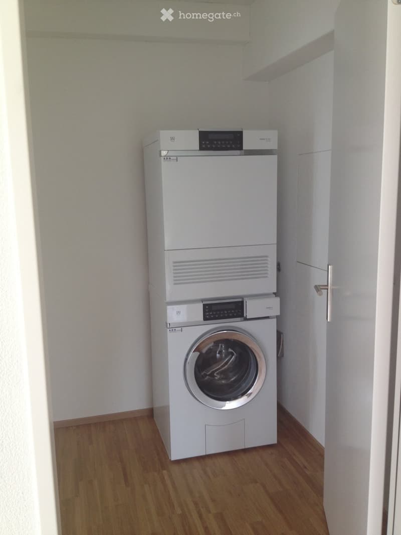 A compact bathroom with a white washing machine and dryer unit against the wall, wooden flooring, and white walls.