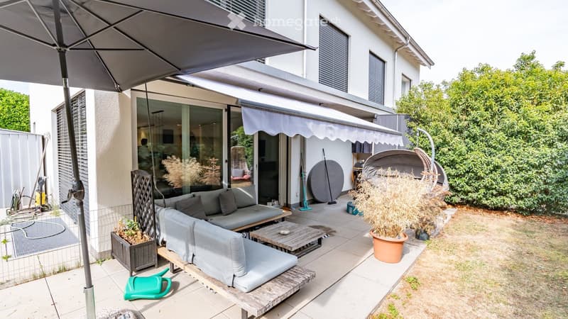 Outdoor patio area with a grey sofa, wooden table, umbrella, wicker chair, and potted plants. The patio is attached to a house with large glass doors.