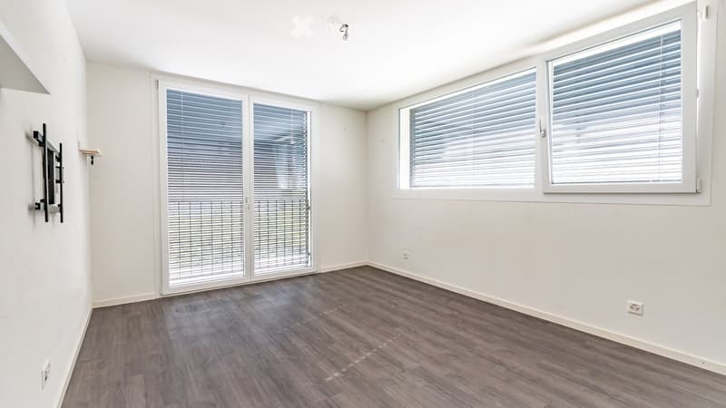 Empty room with wooden flooring, white walls, large windows with blinds, and a sliding glass door leading outside.