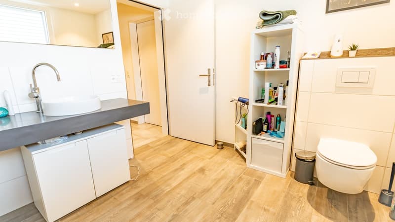 Modern bathroom with a white sink, chrome faucet, white toilet, wooden floor, white cabinet, and a shelf with toiletries and towels.