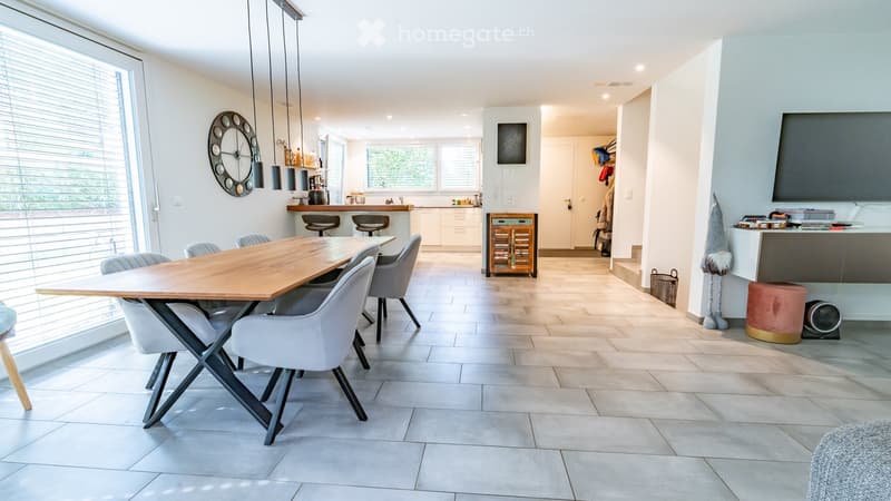 Open-plan dining and kitchen area with a wooden dining table, modern chairs, a kitchen island with bar stools, and a large wall-mounted screen.