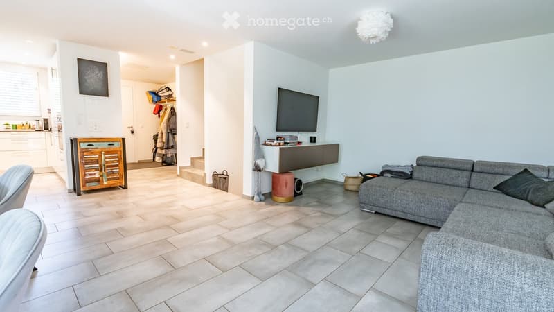 Modern living room with a grey sectional sofa, coffee table, and a TV mounted on the wall. The space features light-colored tiles and a cozy, open layout with a small cabinet and a chair.