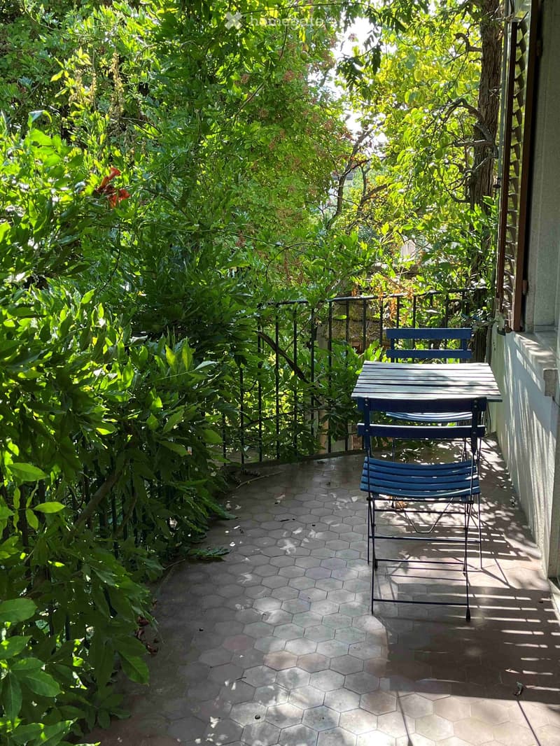 A small, enclosed balcony with a hexagonal tile floor, surrounded by lush greenery and a metal railing. A blue folding chair and a matching table are placed on the balcony.