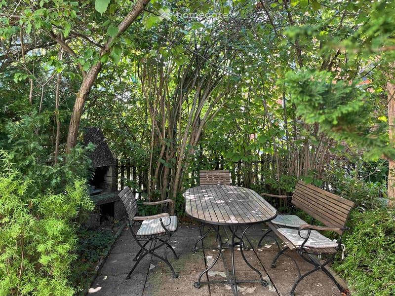 A small outdoor seating area with a wooden table and two matching chairs, surrounded by lush greenery and trees, with a small stone barbecue in the background.