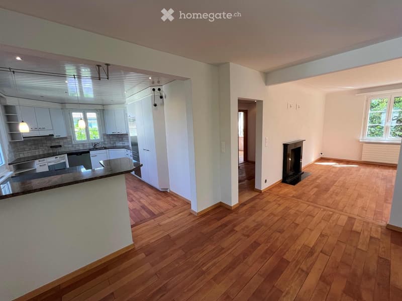 Open-plan living and kitchen area with wooden flooring, white kitchen cabinets, a central island, and a fireplace.