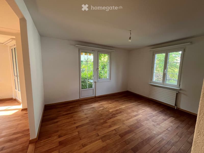 Empty room with wooden flooring, white walls, and large windows allowing natural light. Features a radiator and a door leading to another space.