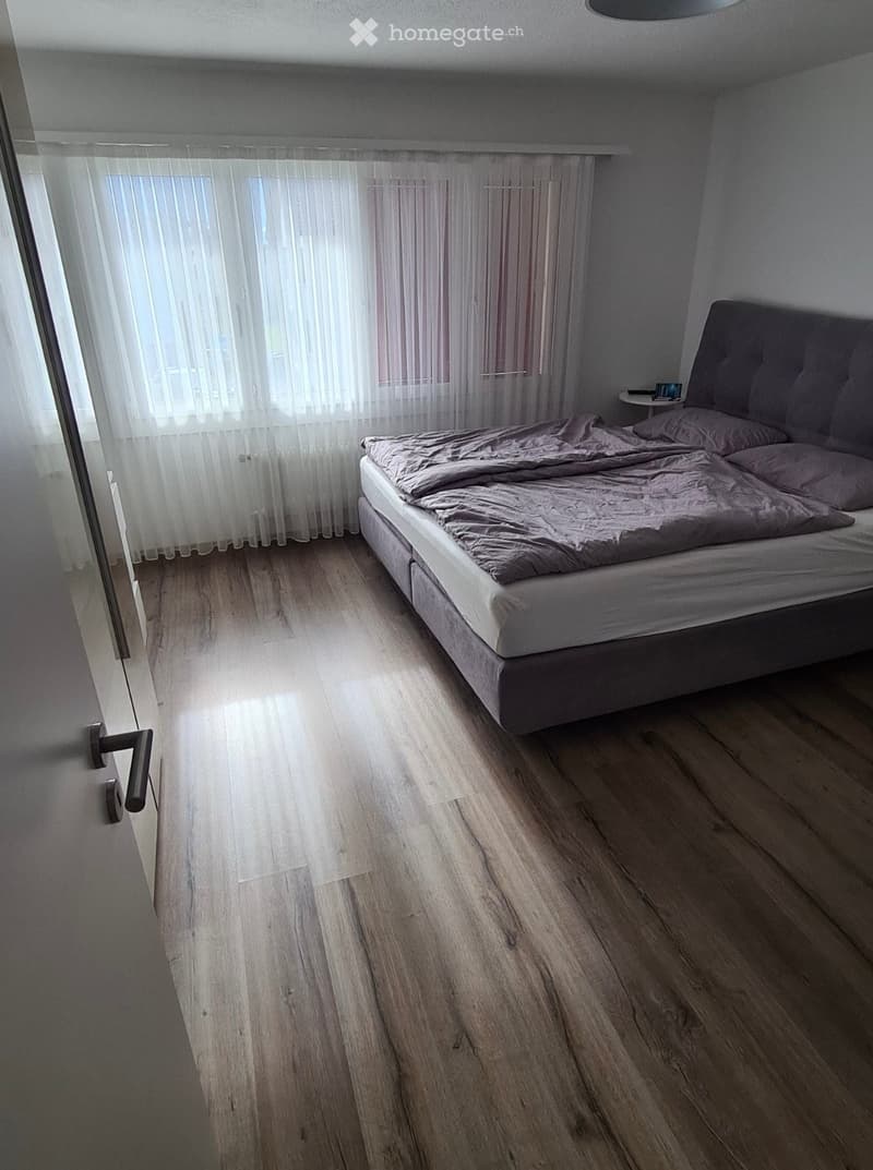 Bedroom with wooden flooring, a bed with a grey duvet and pillows, a small nightstand with a tablet, and sheer white curtains covering a window.