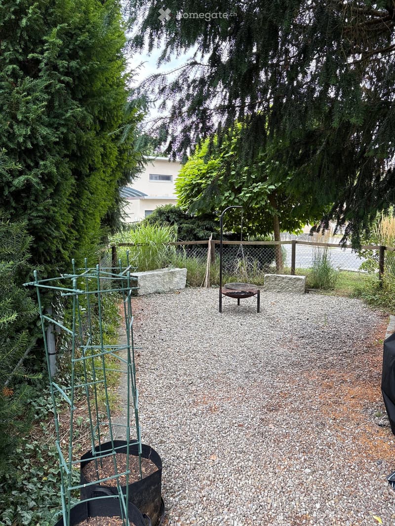 Garden area with gravel pathway, metal trellis, stone planters, and a small metal fire pit surrounded by trees and greenery.