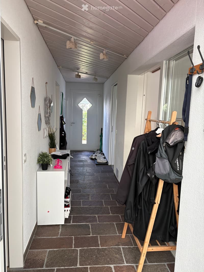 A well-lit hallway with a tiled floor, wooden coat rack, small potted plants, and a white cabinet with shoes and bags.