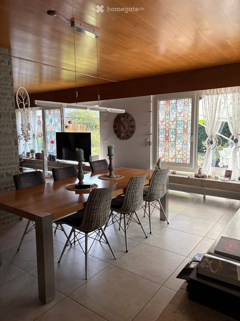 Bright dining area with a wooden dining table surrounded by wicker and black chairs, a television, candle holders, a wall clock, and books on a side table.