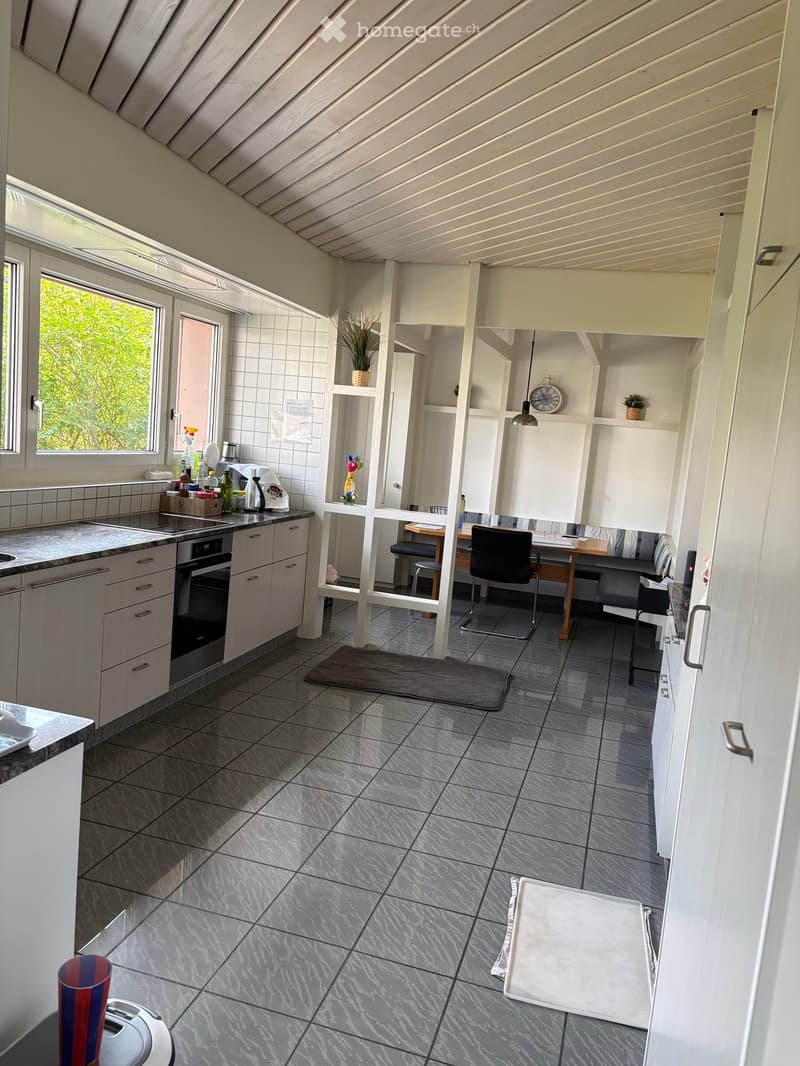 Modern kitchen with white cabinets, grey tiled floor, sink, oven, and a small dining area with chairs and a table.
