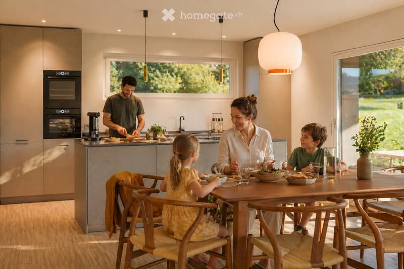 Modern kitchen with a wooden dining table and chairs, a pendant light, and a family enjoying a meal. The kitchen features an oven, a coffee maker, and a plant on the counter.