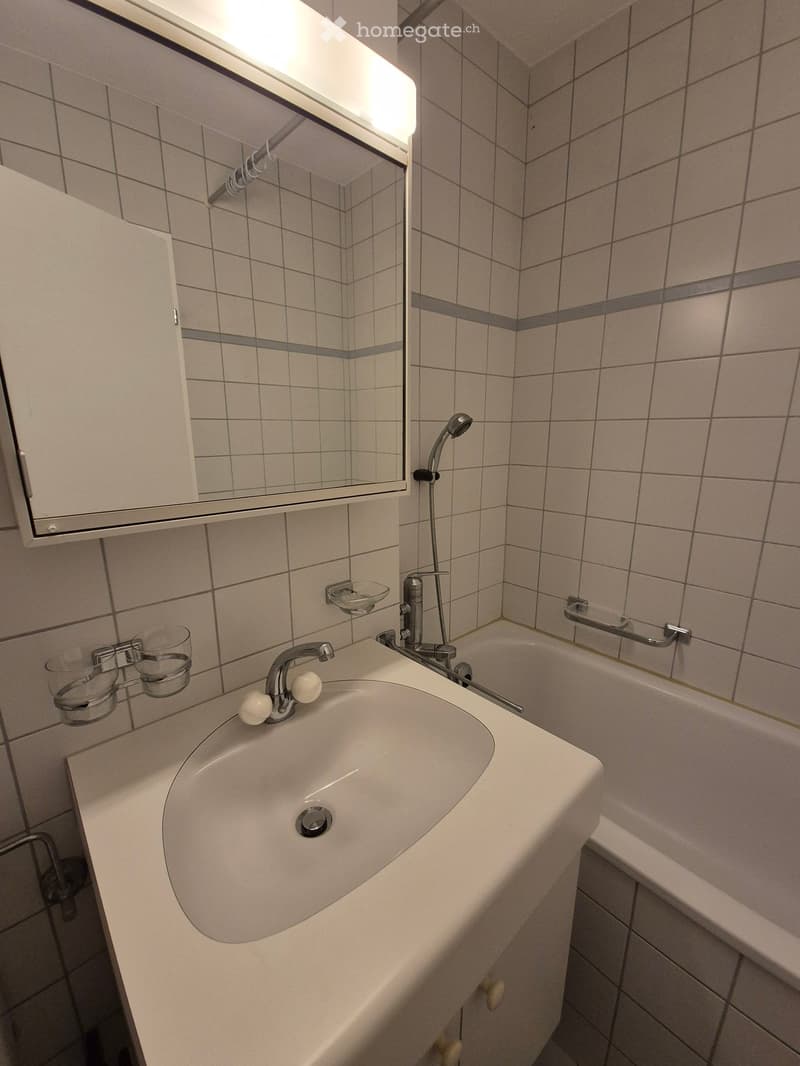 A modern bathroom with white tiled walls and floor, equipped with a sink, a mirror, a bathtub, a shower head, and a faucet. Under the mirror, there are three glass cup holders.