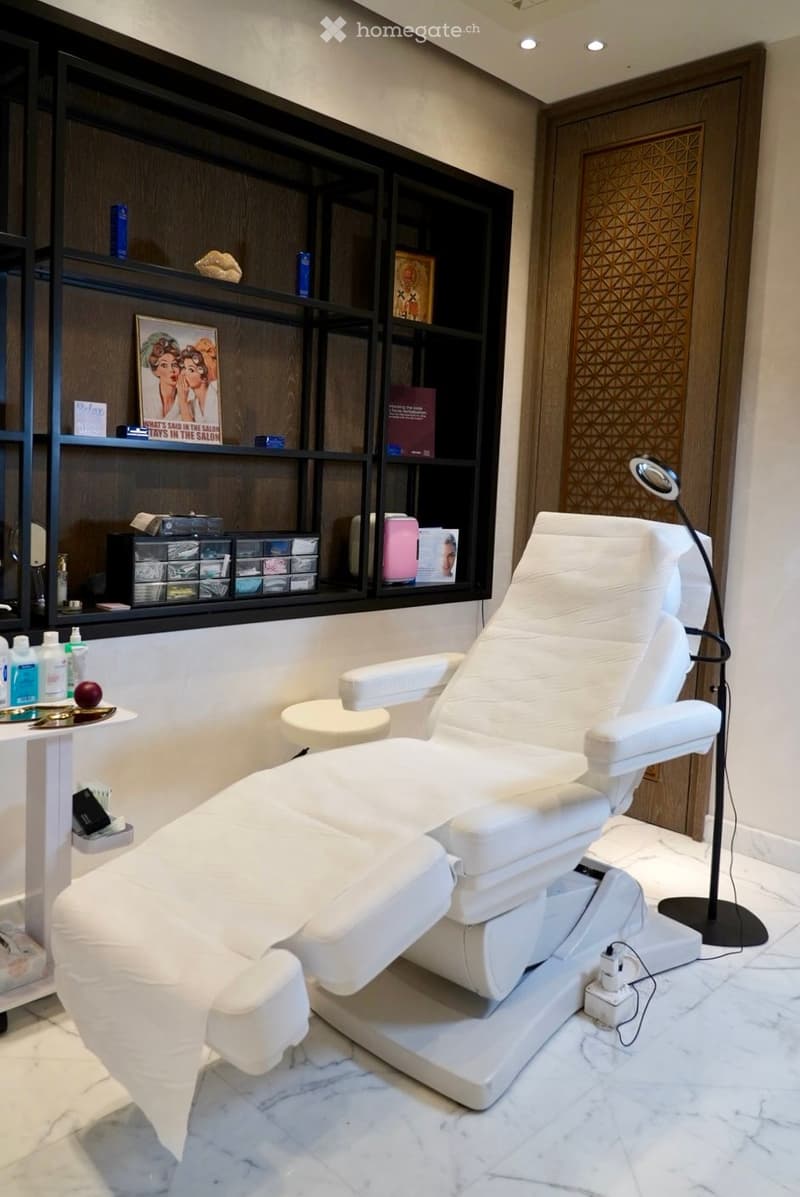 A modern, well-lit salon or spa room with a white reclining chair, a small table with various items, shelves with books and products, and a decorative mirror on the wall.