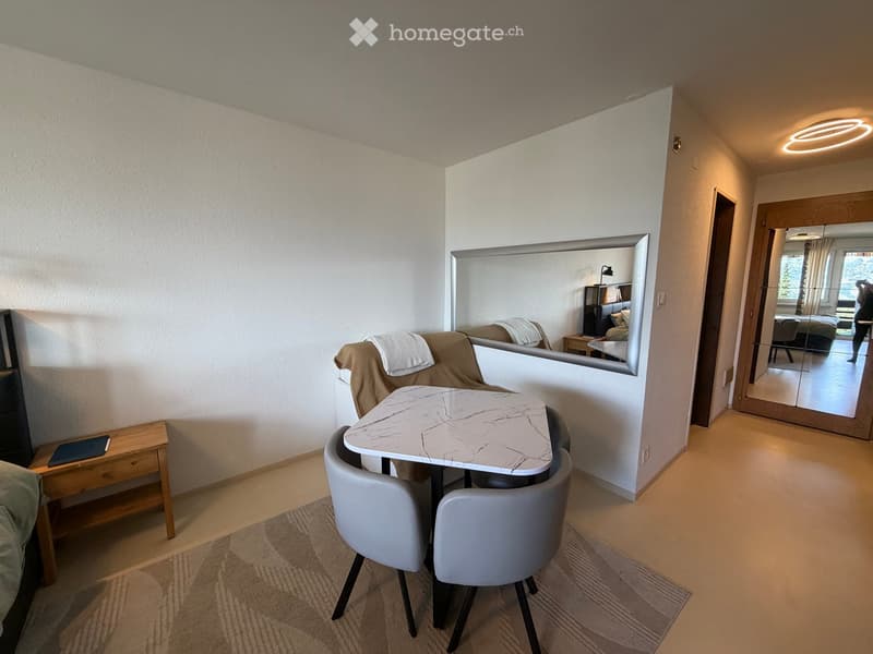 Modern, well-lit living space with a sofa, small dining table with chairs, a mirror, and a nightstand with a lamp visible in the adjacent bedroom.
