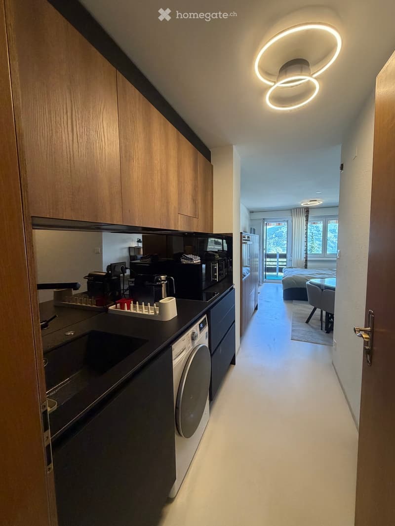 Modern hallway with sleek wooden cabinets, integrated kitchen area visible, leading to a bedroom with a bed, table, and chairs.