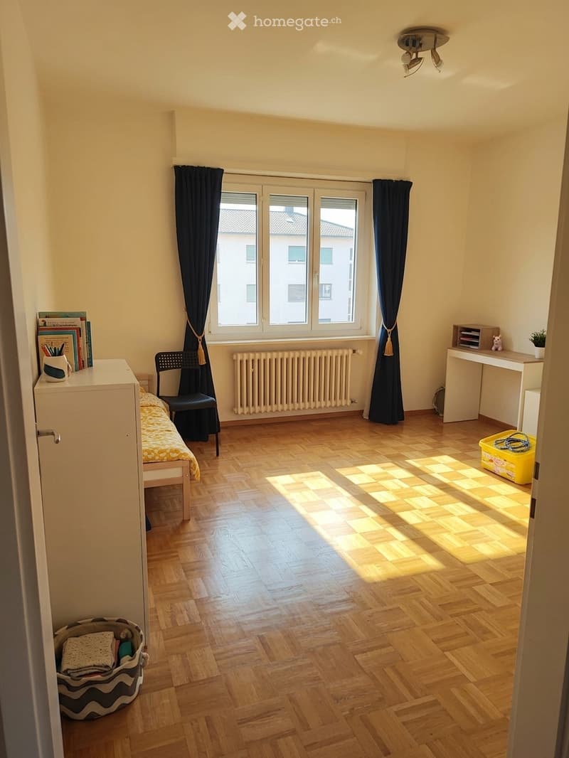 Bright bedroom with wooden flooring, large window with dark curtains, bed with yellow bedding, chair, desk, radiator, and small storage items.