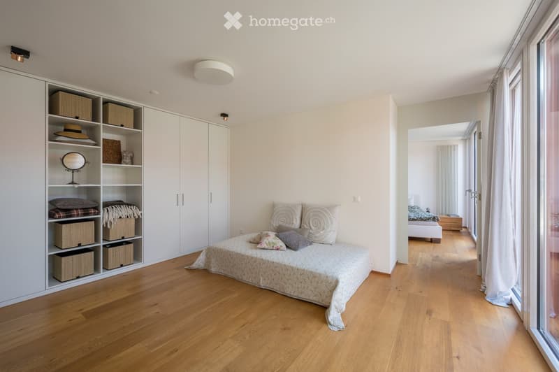 A bedroom with a bed, pillows, and a blanket. There is a white wardrobe with shelves and baskets. The room has a wooden floor and white walls. There is a glass door leading to another room.