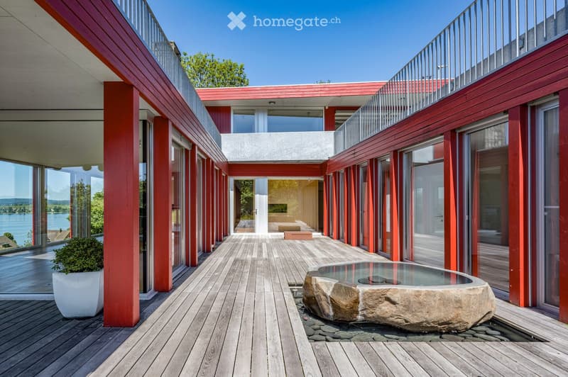 modern building with red exterior, wooden deck, large windows, water feature, balcony, view of the lake
