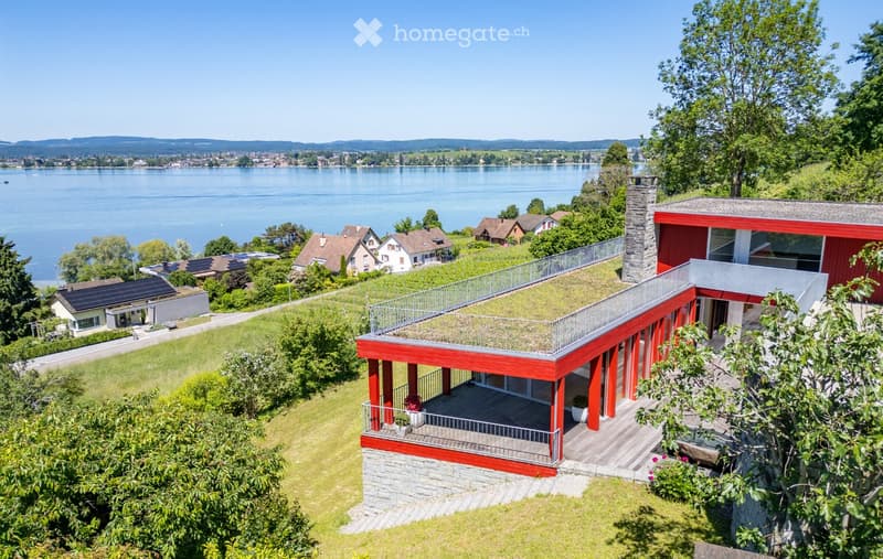 Modern red house with a green roof and balcony overlooking a lake, surrounded by trees and neighboring houses.