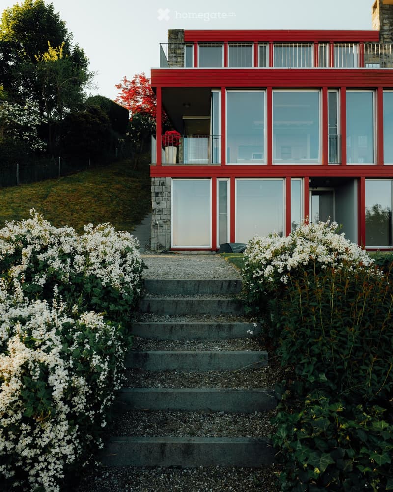 Modern house, red exterior, large glass windows, balcony, steps leading to entrance