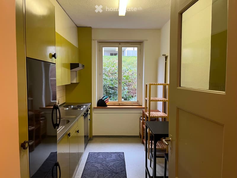 A small, modern kitchen area with light green cabinets, a stove, refrigerator, and a small dining area with a table and chairs. A window offers a view of greenery outside.