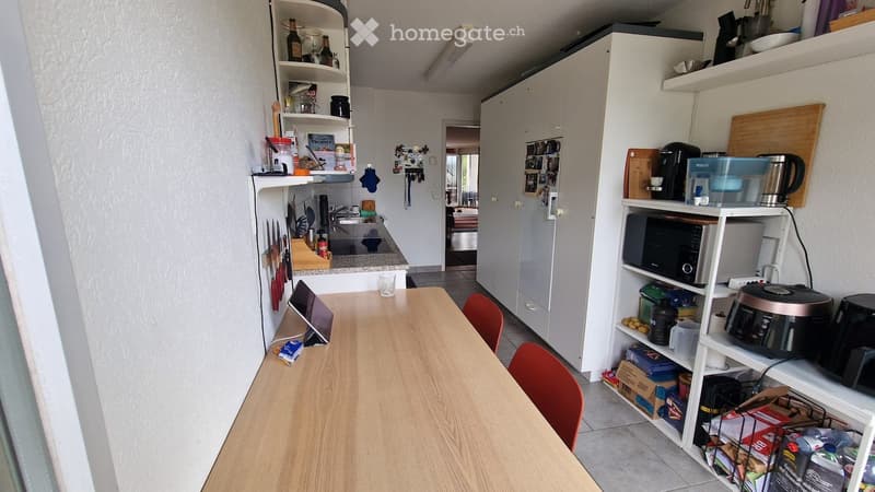 The kitchen features a wooden dining table with red chairs, white cabinets, a refrigerator, microwave, and various kitchen appliances on shelves. There are also bottles and other kitchen items on the shelves.