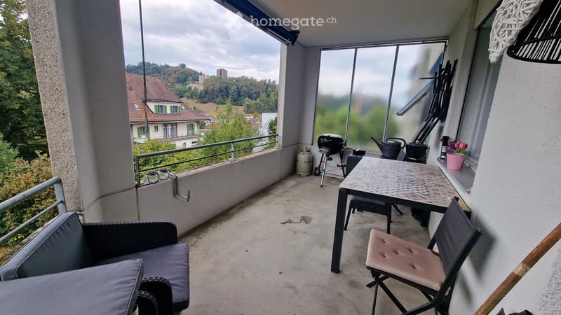 Balcony with a sofa, table, chairs, flower pot, barbecue, and teapot, overlooking a scenic view of a town and greenery.