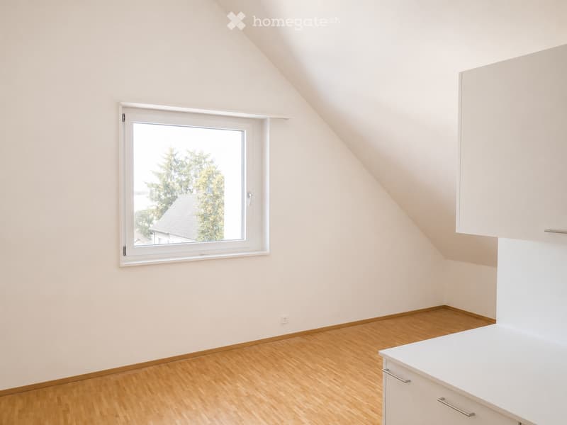 Empty attic room with wooden flooring, white walls, and a large window showing a view of trees and a roof outside.