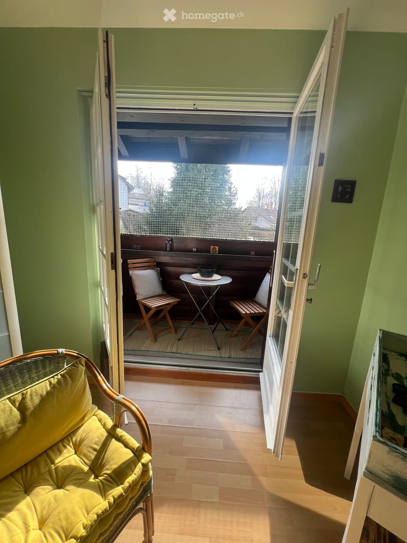 Furnished Room. Bright room with wooden flooring, a yellow sofa, and an open sliding door leading to a small balcony with a round table and two chairs.
