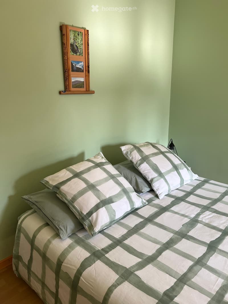 Bedroom. A neatly made bed with a green and white checkered duvet and matching pillows, positioned against a light green wall with a small wooden shelf holding framed pictures.