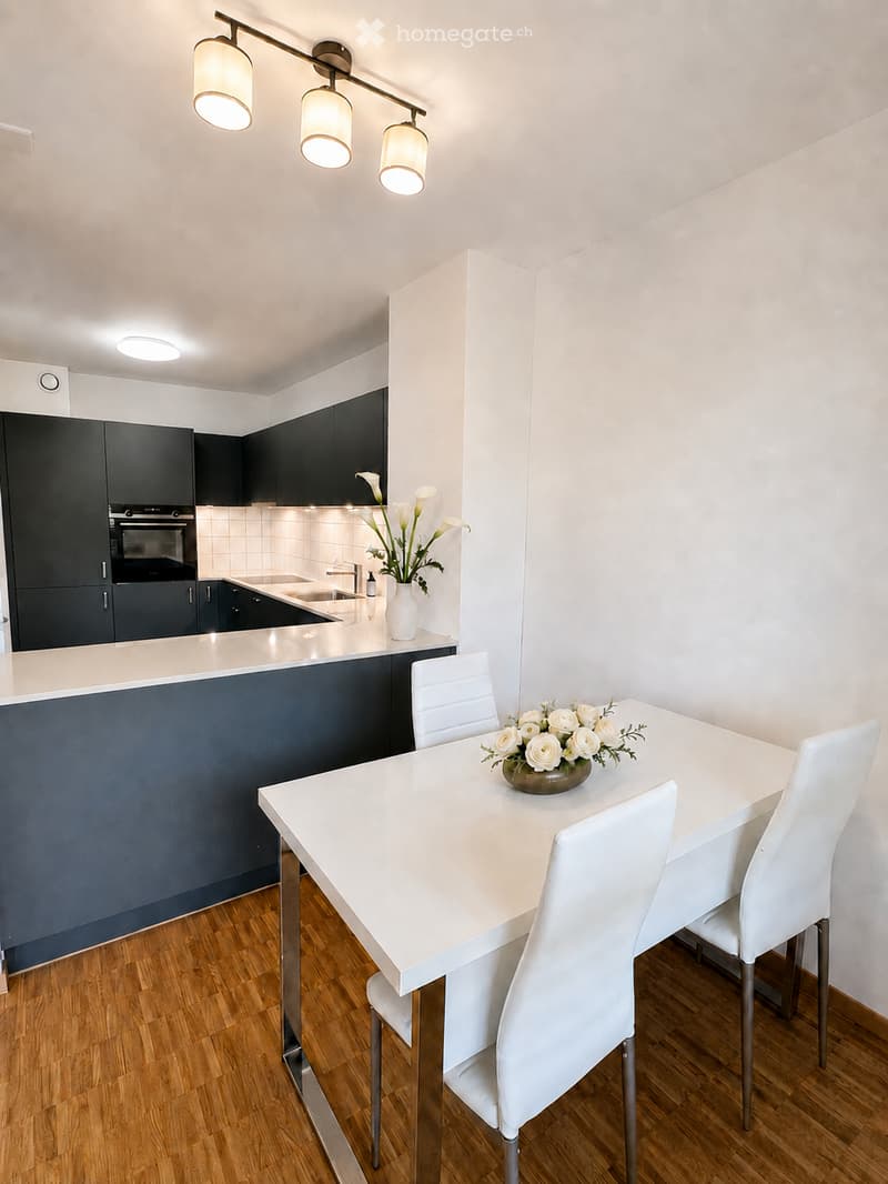 Modern kitchen and dining area with white dining table, four white chairs, dark blue cabinets, stainless steel sink, and decorative flower vases.