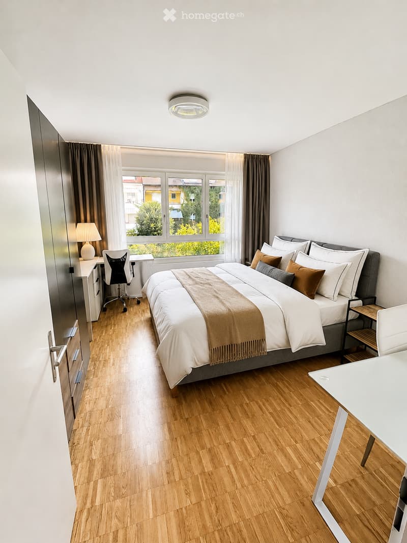 Modern bedroom with a large bed, wooden flooring, a desk with a chair, and a window with curtains.