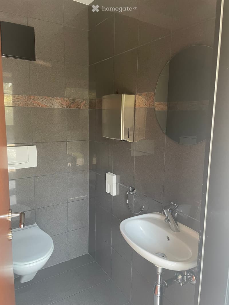 Modern bathroom with a white sink, chrome faucet, toilet, and a round mirror. Grey tiled walls and floor.