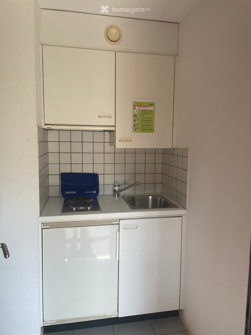 Compact kitchen with white cabinets, a small sink, a single-burner stove, and a refrigerator. The walls are tiled, and there is a notice poster on the wall.