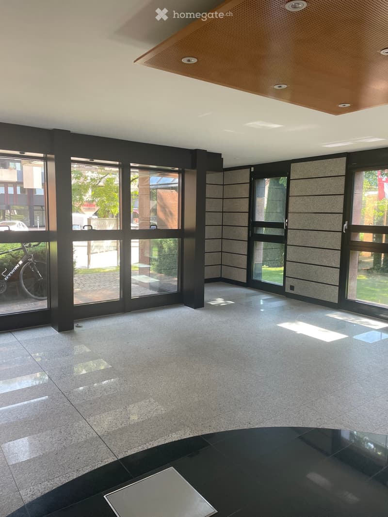 Empty lobby area with polished concrete floors, large glass doors, and a black reception desk. The space is well-lit with recessed lighting and features a modern, minimalist design.