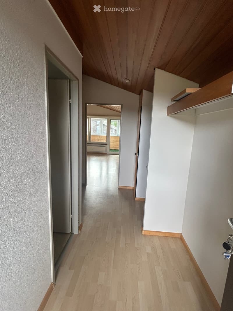 A clean, well-lit hallway with light wooden flooring, white walls, and a wooden ceiling. The hallway features several doors and a small built-in shelf on the right side.