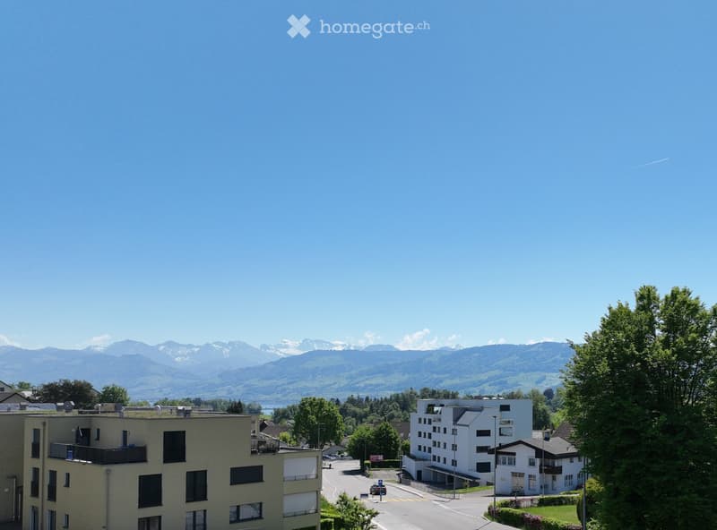 A multi-story residential building with balconies and windows, surrounded by greenery and a clear view of snow-capped mountains in the background.