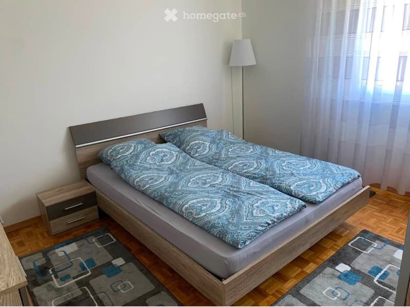 Bedroom with a double bed, nightstand, lamp, and rug. The bed has a wooden headboard and is covered with blue patterned bedding.
