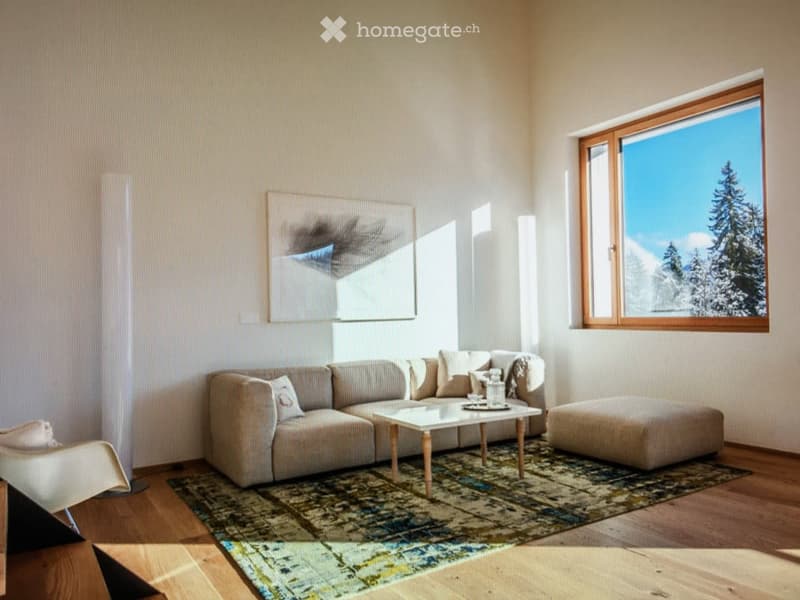 Bright living room with large windows offering a scenic view, featuring a beige sectional sofa, a glass coffee table, an ottoman, a white armchair, and a patterned rug.