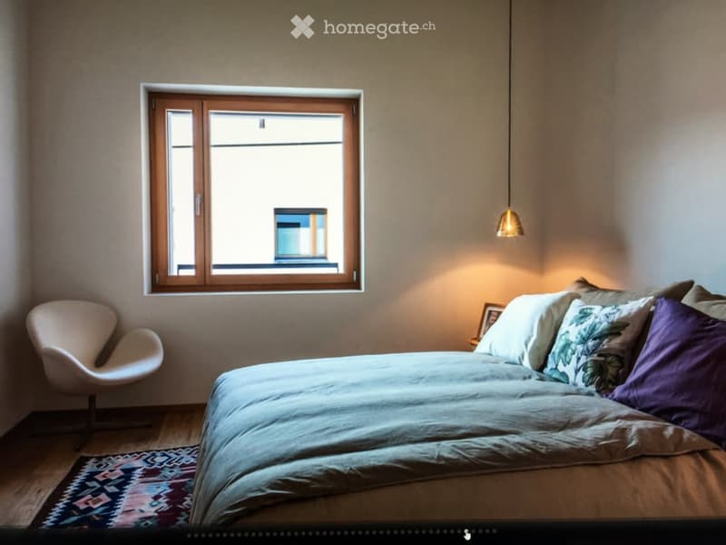 A modern bedroom with a large window, a neatly made bed with pillows, a white chair, a patterned rug, and a hanging pendant light.