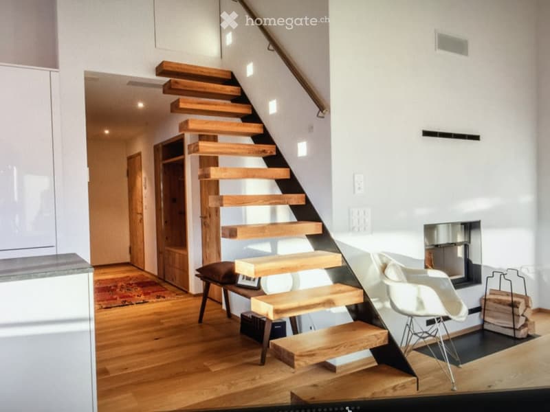 Modern interior with a wooden staircase, a white chair, a small stool, and a fireplace in the background.