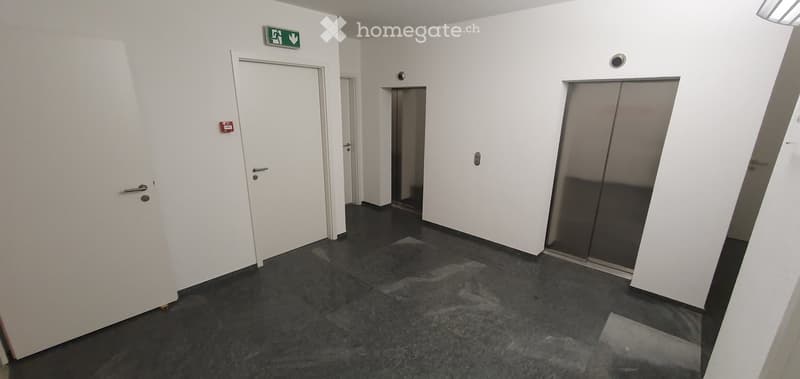 Corridor with 2 doors, 2 elevators, gray tiles, white walls, emergency exit sign