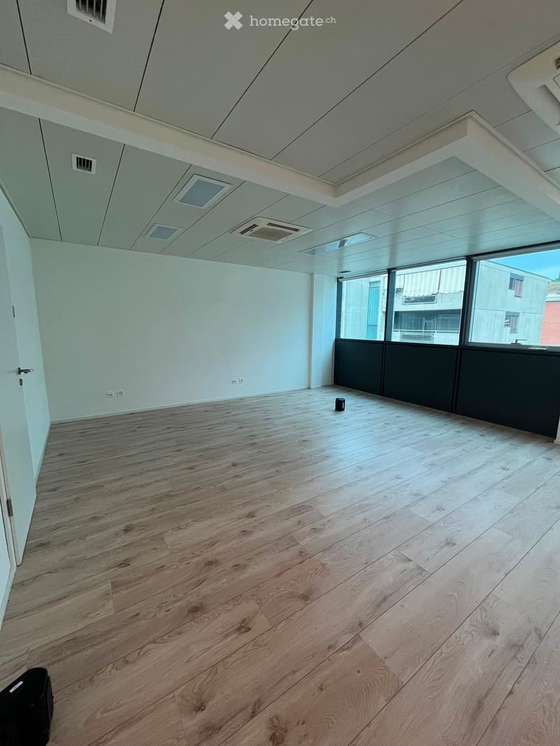 Empty room with wooden flooring, white walls, and large windows offering a view of the outside. The ceiling has several vents and a skylight.